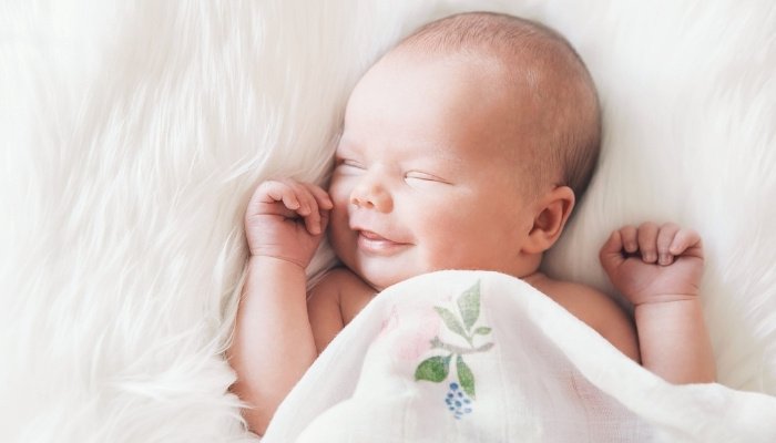 A newborn baby sleeping under a white blanket with a smile on his face.