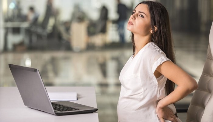 A young pregnant woman holding her back as if in pain while at work.