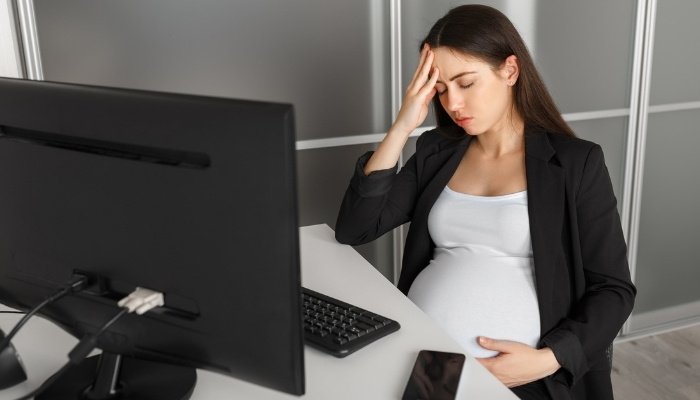 A pregnant woman sitting in her office at work looking tired, sick, and stressed.