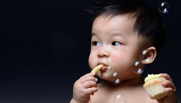 A cute baby boy eating a French fry with one hand and holding a cupcake in the other.