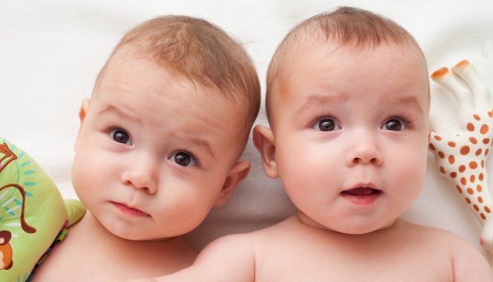 Baby twin brothers with no shirts on and toys lying near their heads.