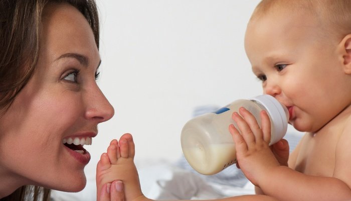 A smiling mother playing with her baby's foot while he drinks a bottle.