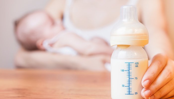 A full baby bottle in the foreground with a nursing baby visible in the background.