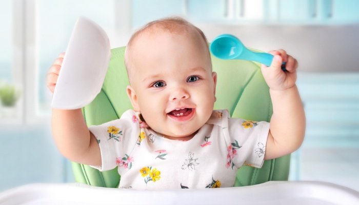 A cute baby in a high chair holding up a bowl and a spoon in her hands.