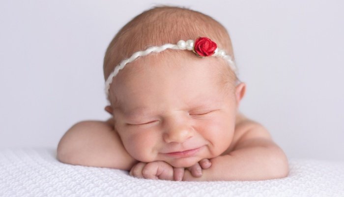 A newborn baby girl wearing a rose headband with her head propped on her hands.