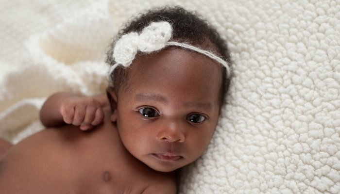 A newborn baby girl wearing a white bow on her head and lying on a faux sheepskin blanket.