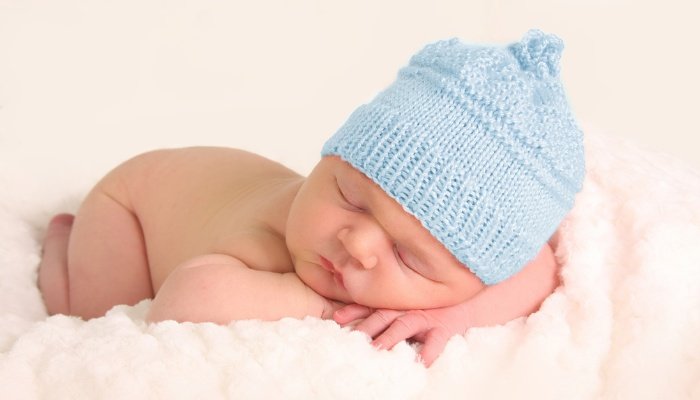 A newborn baby boy lying on his stomach, naked except for a blue hat.