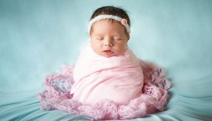 A cute newborn baby girl swaddled snugly and sitting upright in a nest of blankets.