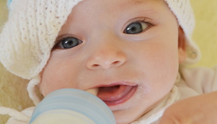 A cute baby wearing a knit hat, smiling, and playing with the bottle nipple instead of drinking.