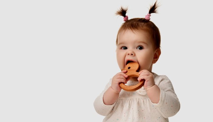 A baby girl with pigtails on top of her head chews on a wooden teether.