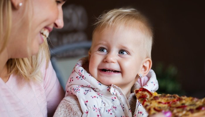 A mother smiles while offering her baby a bite of pizza.