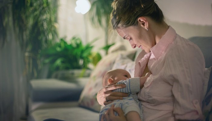 A mother seated in a room with plants and soft light breastfeeding her baby.