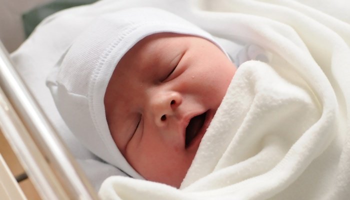 A newborn baby in a hat sleeping in a hospital bassinet.