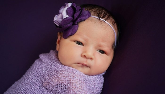 A newborn baby girl wrapped in a purple blanket and wearing a purple rose headband.