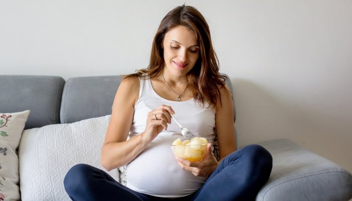 A pregnant woman sitting on a couch eating sliced fruit from a bowl.