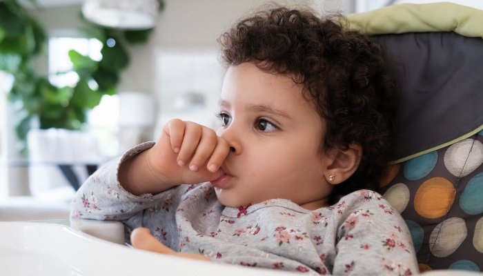 A toddler with curly hair sucking her thumb while sitting in her high chair.