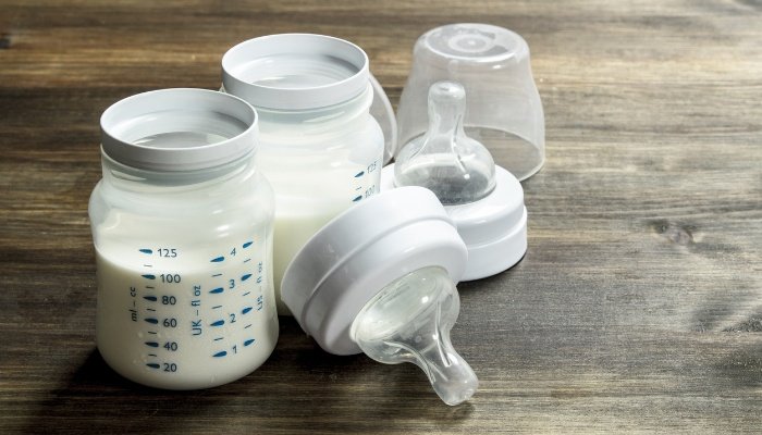 Two small baby bottles full of milk beside the nipples and caps on a wood table.