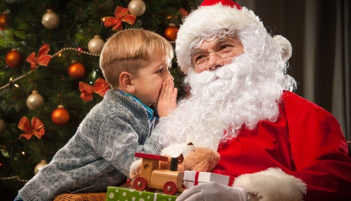 A little boy whispering in Santa's ear in front of a Christmas tree.