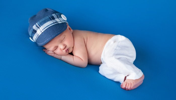 A newborn baby boy wearing only pants and a hat sleeping on his side against a blue background.