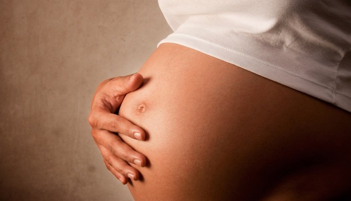 A woman with her hand on her pregnant belly against a beige background.