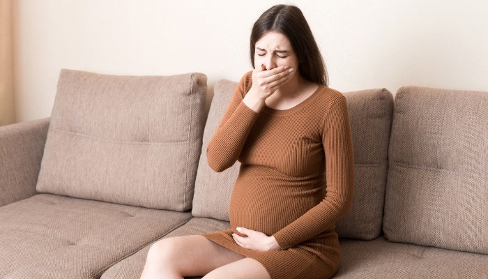 A pregnant woman sits on the couch covering her mouth because she feels nauseous.