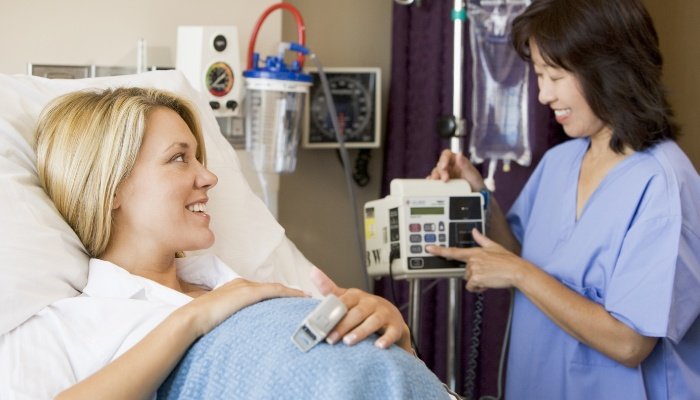 A smiling pregnant woman converses with her nurse while lying in a hospital bed.