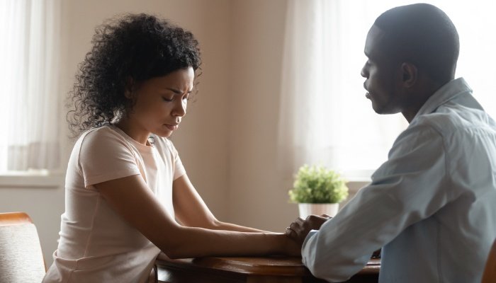 A couple sitting together at a table holding hands and looking upset.