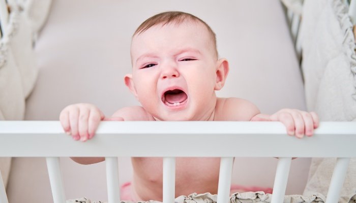 A baby boy standing in his crib, clinging to the rail and crying.