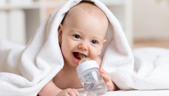 A baby with a bath towel on his head drinking water from his baby bottle.