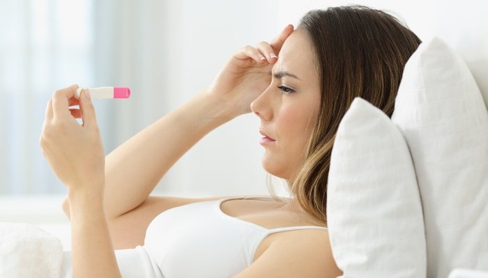 A woman lying in bed staring at a pregnancy test and looking confused.