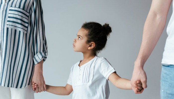 A little girl standing in between her divorced parents, holding hands with both of them.