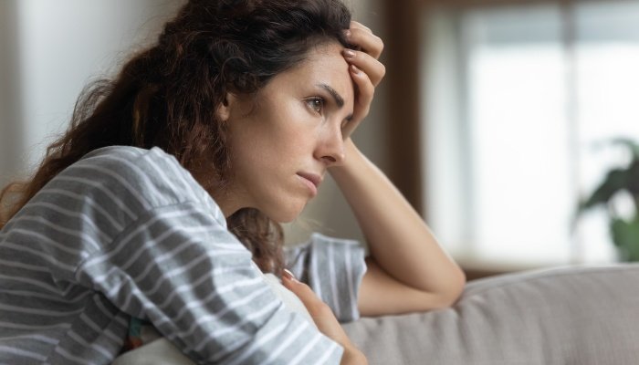 A sad-looking woman with her head resting on her hand while leaning against the back of the couch.