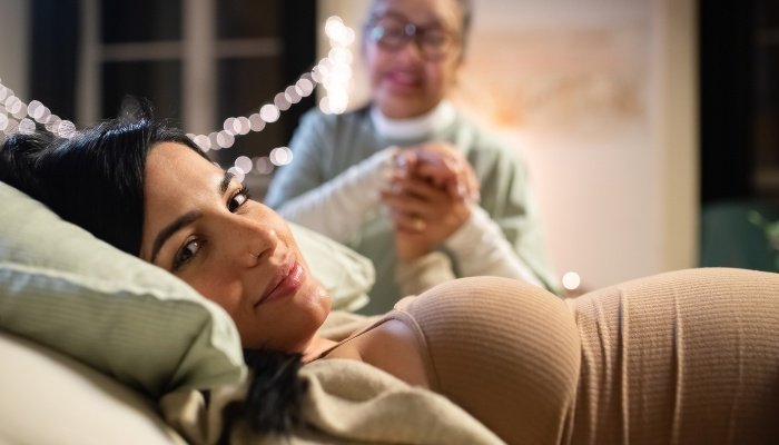A smiling pregnant woman in bed with her doula in the background holding her hand.
