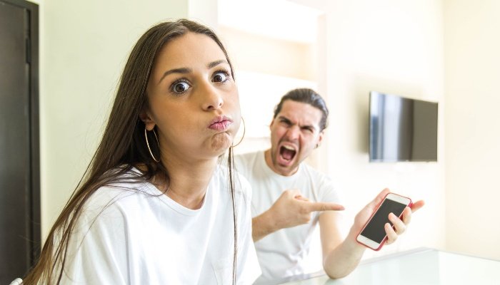 A woman looking stressed with her boyfriend screaming in the background and pointing to a phone.