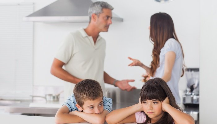 Two sad kids sit at the kitchen table while listening to their parents argue behind them.