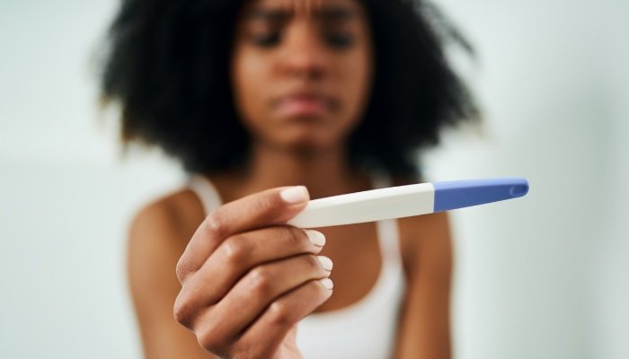 A woman in a white tank top holding up a pregnancy test to examine it.