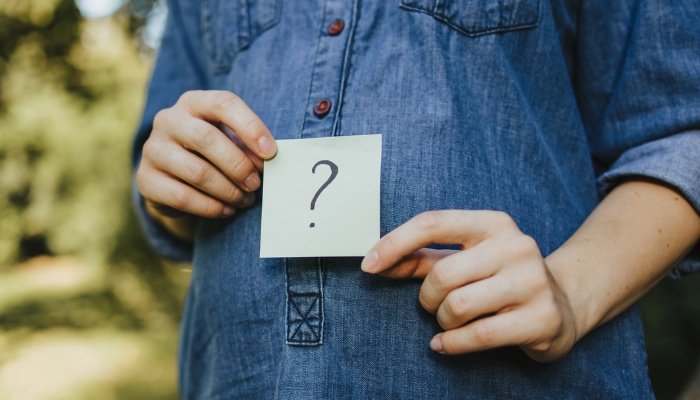 A pregnant woman in a denim dress holding a question mark sign over her belly.