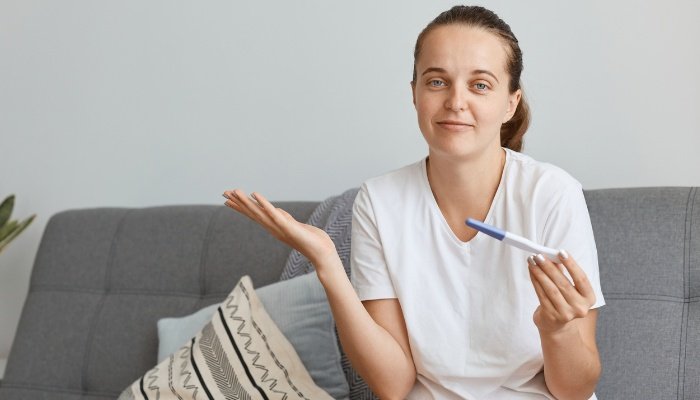 A young woman sitting on her couch with a pregnancy test and looking confused.