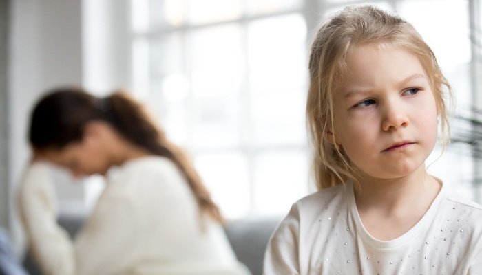 An angry little girl in the foreground with a frustrated woman holding her head in the background.