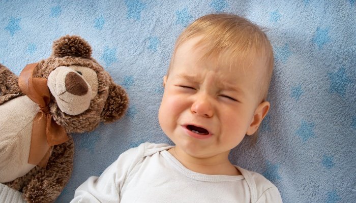 A baby boy lying in bed beside a teddy fussing and crying.