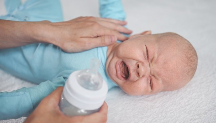 A baby boy lying on a bed crying while his mother places her hand on his chest and holds a bottle.