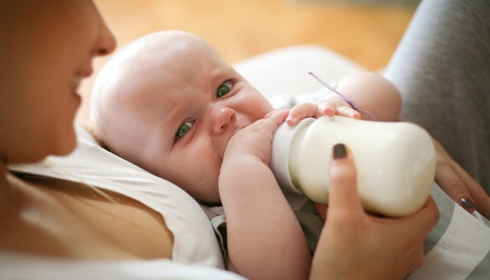 A baby looking very unhappy with the bottle offered him by his mother.