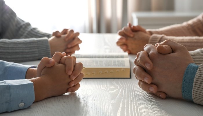 The hands of two boys and their godparents folded for prayer at a kitchen table.