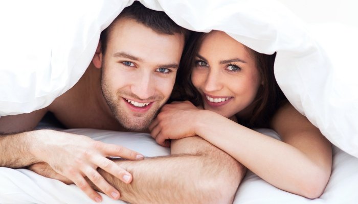 A young couple peeking out from under the covers in bed.