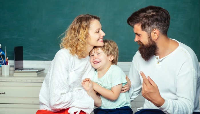 A mother and father lovingly snuggle their young son in a bedroom with dark-green walls.