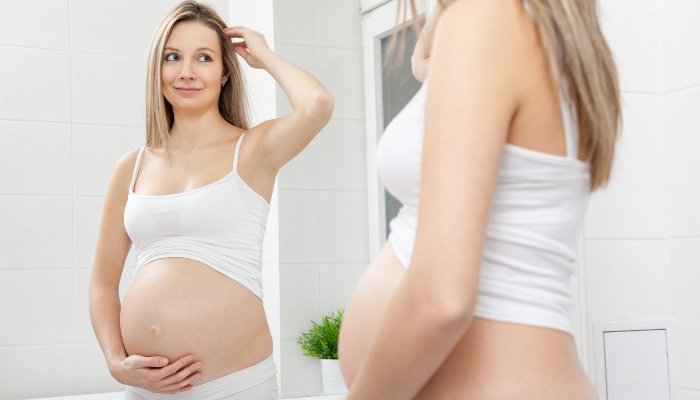 A pregnant woman staring at her belly in the bathroom mirror and smiling.