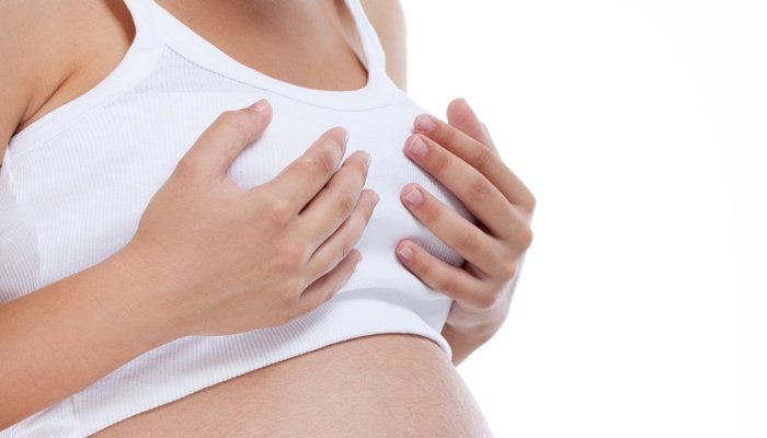 A pregnant woman in white tank top holding her breasts.