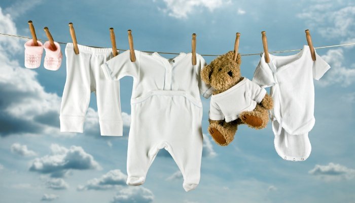 White baby clothes and a teddy bear hanging on a clothesline under a blue sky.