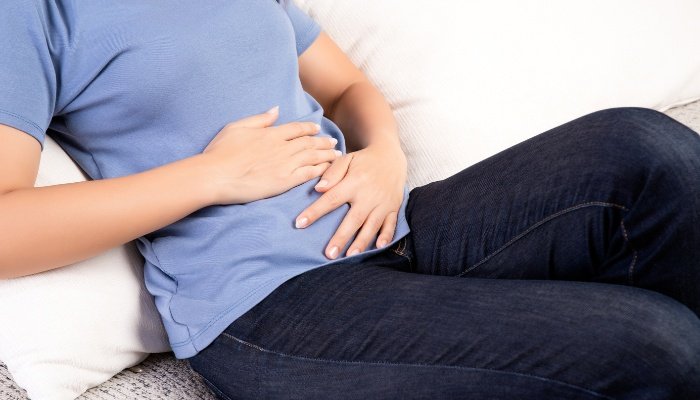 A woman sitting on a white couch holding her stomach and feeling bloated.