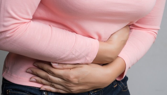 A woman wearing a pink top and jeans holding her bloated belly as if in pain.